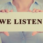 Closeup retro vintage image businesswoman, female, girl, person hands holding white rectangle sign or card with message WE LISTEN isolated on gray office wall background. Counseling concept